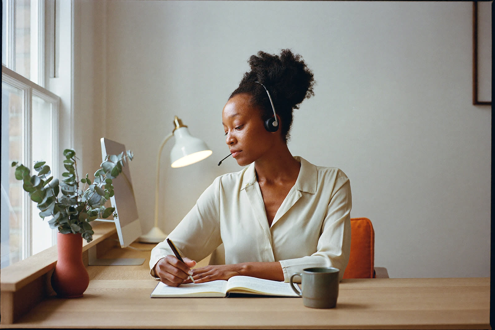 A receptionist at a bright front desk, wearing a headset, writing an enquiry into an appointment book.